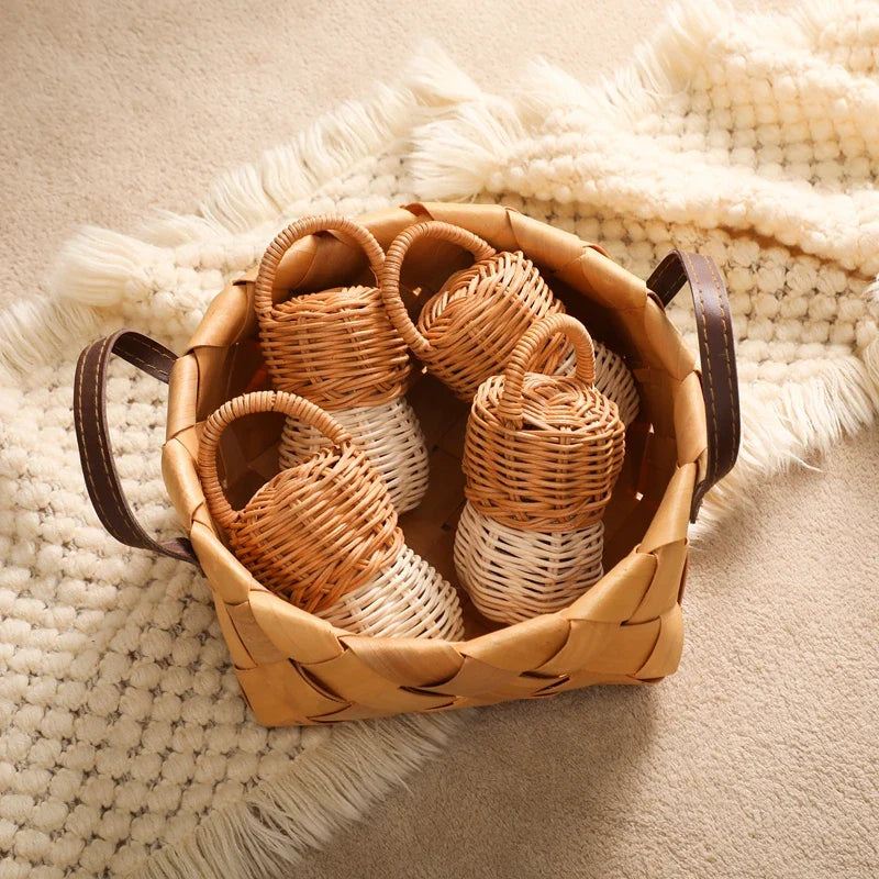 Ein geflochtener Korb mit dunklen Griffen enthält fünf kleine, zweifarbige Weidenkörbe und den Hello World Rassel Eichel [6x13,5cm], ein charmantes Naturspielzeug. Das Arrangement steht auf einer cremefarbenen, strukturierten Decke auf einem beigen Teppich.