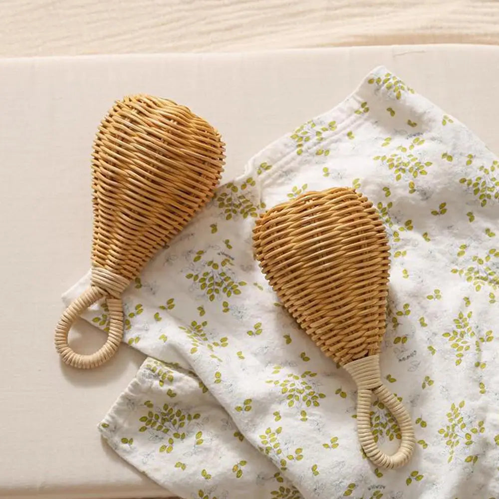 Zwei Rassel Rattan [8x17cm] von Hello World mit geschwungenen Griffen liegen auf einem weißen Tuch mit kleinen grünen Blättern und heben sich sanft vom hellen Hintergrund ab.