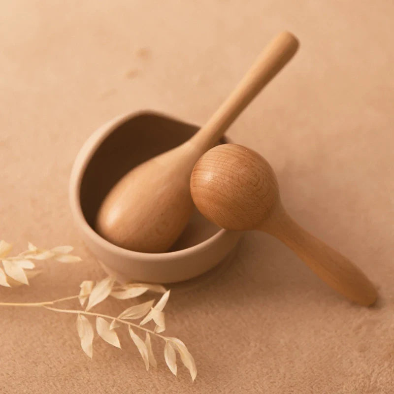 Die Hello World Rassel gross [5x19,5cm] aus Holz liegt auf einer weichen, beigen Fläche-eine im Schälchen, eine daneben. Getrocknete Deko-Blätter sorgen für Charme. Das perfekte erste Musikinstrument für Kinder!.