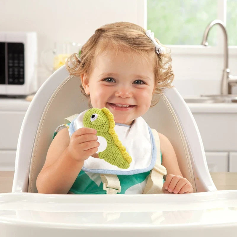 Ein lächelndes Kleinkind sitzt mit einem Lätzchen in einem Hochstuhl und hält den Hello World Rasseltier Dino [10x14cm], ein grün-gelbes gehäkeltes Dinosaurier-Spielzeug, in einer hellen Küche.