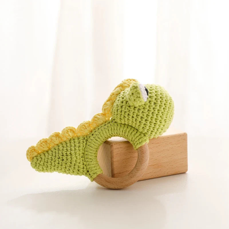 Das Hello World Rasseltier Dino [10x14cm], eine handgehäkelte grüne Babyrassel mit einem hölzernen Ringgriff, steht auf einem kleinen Holzklotz auf einer hellen Fläche mit einem weichen weißen Hintergrund.