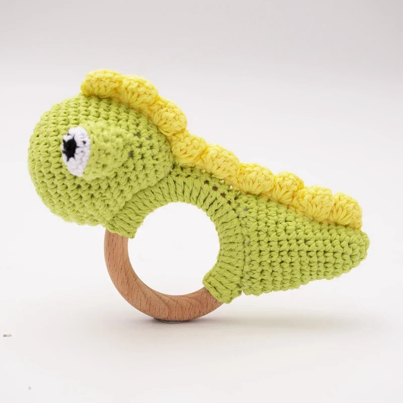 Das Hello World Rasseltier Dino [10x14cm] ist eine grüne gehäkelte Dinosaurier-Rassel mit gelben Stacheln auf einem Holzring, der auf einem schlichten weißen Hintergrund abgebildet ist.