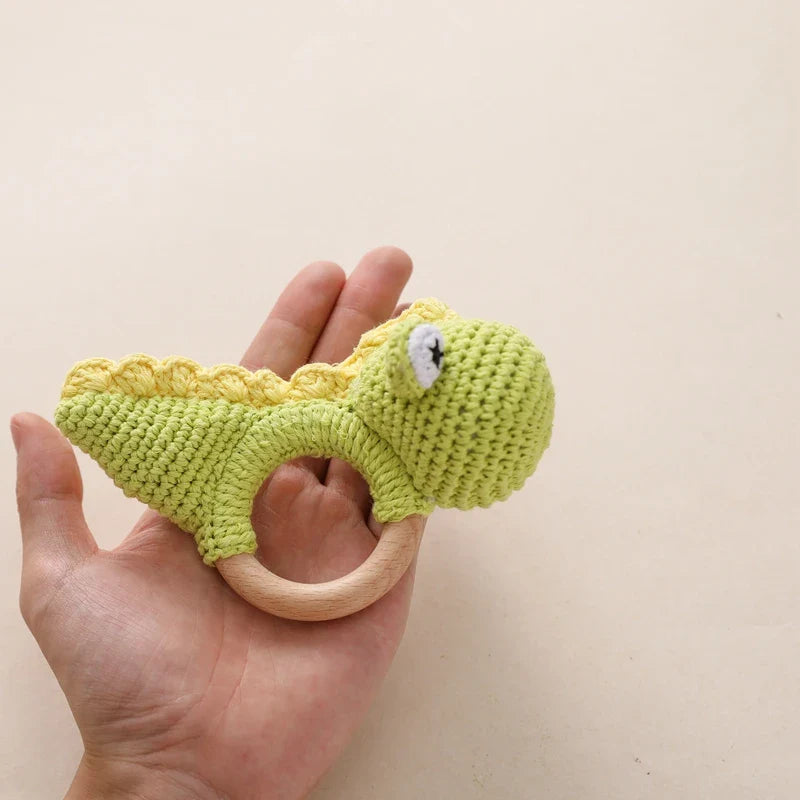 Eine Hand hält den Hello World Rasseltier Dino [10x14cm] mit einem Holzring vor einem beigen Hintergrund. Das Spielzeug zeigt einen grünen, rundköpfigen Dinosaurier mit einem kleinen Auge und einem strukturierten gelben Rücken.