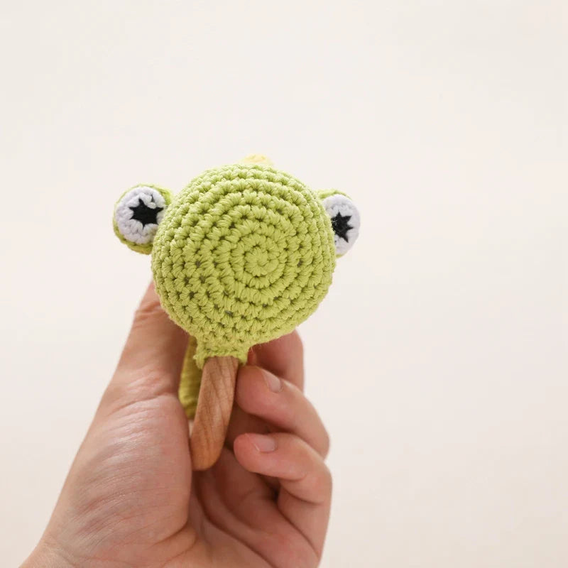 Eine Hand hält das Rasseltier Dino [10x14cm] von Hello World - eine grüne, handgefertigte Dino-Rassel mit runden Augen und einem Holzring - vor einem schlichten hellen Hintergrund.