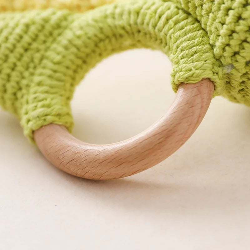 Nahaufnahme eines hellen Holzrings, der an strukturiertem, gelb-grünem Häkelwerk befestigt ist, Teil des Hello World Rasseltier Dino [10x14cm] Accessoires.