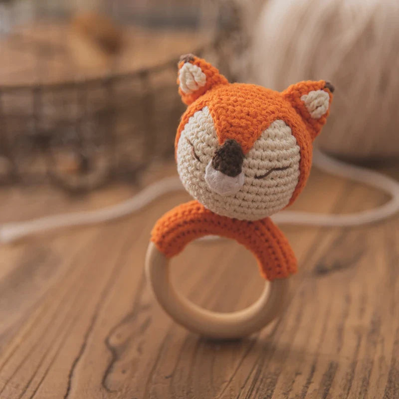Das Rasseltier Fuchs [6.5x12cm] von Hello World mit geschlossenen Augen ist an einem Holzring befestigt und liegt auf einer Holzoberfläche - ein liebevoll handgefertigtes Babyspielzeug, im Hintergrund Garn und Korb unscharf.