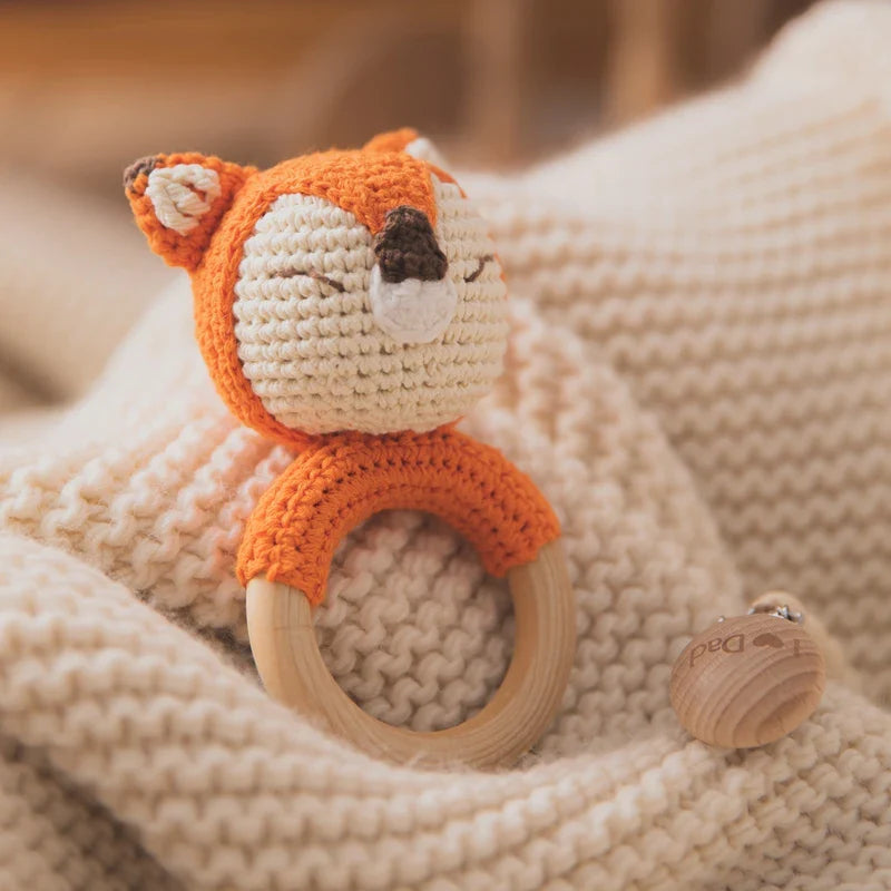 Das handgefertigte Rasseltier Fuchs [6.5x12cm] von Hello World mit Holzring liegt auf einer weichen, beigefarbenen Strickdecke. In der Nähe befindet sich ein weiteres Babyspielzeug Wald mit kleiner Glocke.