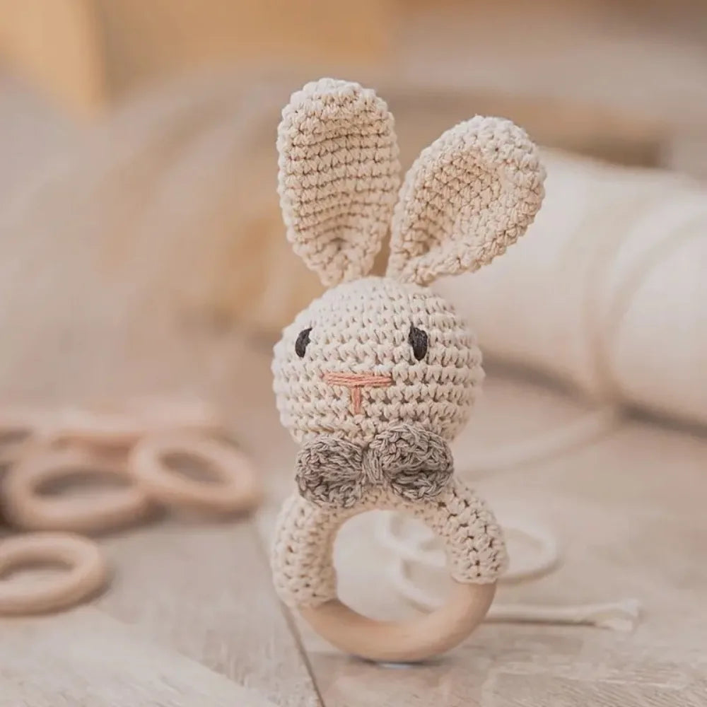 Der handgefertigte Rasseltier Hase [6x15cm] von Hello World mit Holzring, beigem Garnkörper, langen Ohren, schwarzen gestickten Augen, rosa Nase und brauner Fliege liegt auf einer Holzoberfläche.