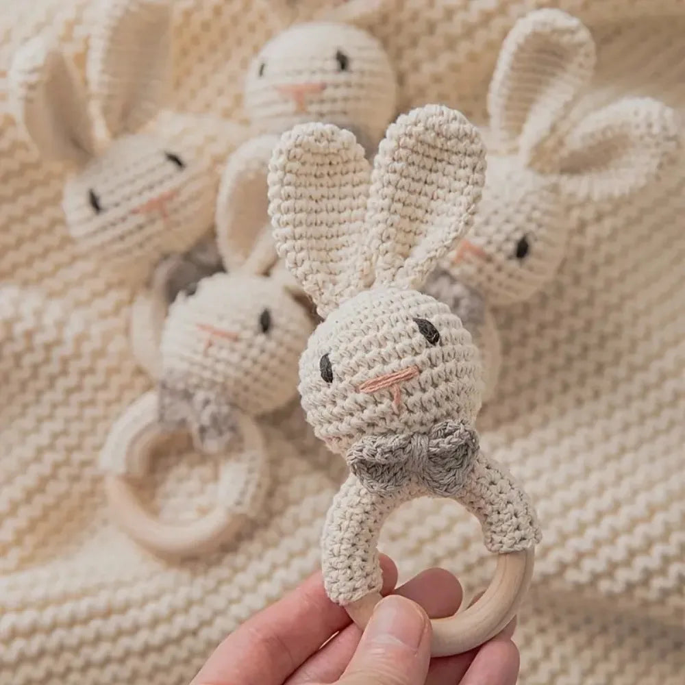 Eine Hand hält einen Hello World Rasseltier Hase [6x15cm] mit langen Ohren und grauer Fliege. Andere ähnliche gehäkelte Hasenrasseln sind im Hintergrund verschwommen, alle auf einer gestrickten beigen Decke.