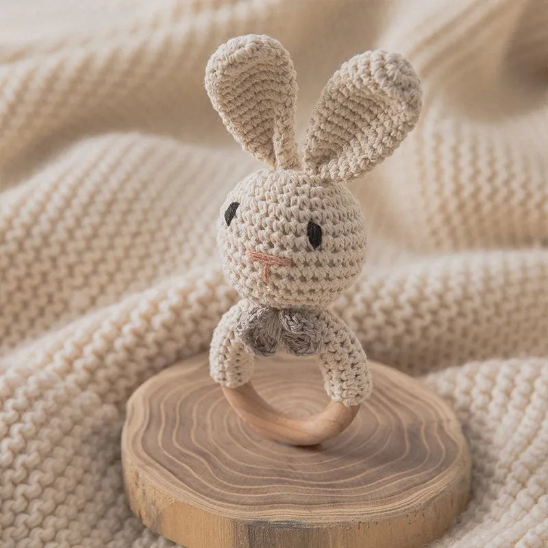 Das handgefertigte Rasseltier Hase [6x15cm] von Hello World mit langen Ohren und Holzring sitzt auf einer Holzplatte, umgeben von einer weichen, beigefarbenen Strickdecke.