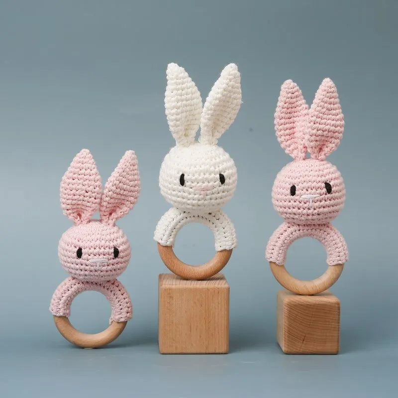 Drei Hello World Rasseltier Hase Rosa [6x15cm] Babyrasseln mit Holzringen stehen auf Holzklötzen vor blauem Hintergrund. Die rosa und weißen Motive sind mit gestickten Augen und langen Ohren versehen - ideal als Prinzessinnen Geschenk.