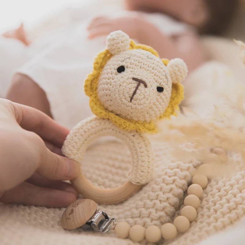 Eine Hand hält das Rasseltier Löwe [7.5x11cm] von Hello World mit Holzring, während ein Baby in weiß auf einer cremefarbenen Strickdecke liegt. Im Hintergrund ist außerdem ein Perlen-Schnullerclip zu sehen.