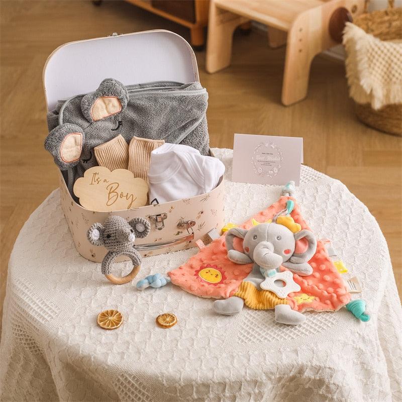 Das Ringo Baby Geschenk zur Geburt von Hello World enthält Kleidung, graue Booties, einen Rasselelefanten, eine Elefanten-Schnuffeldecke, eine Karte und getrocknete Orangenscheiben. Im Hintergrund stehen ein Holzspielzeug und ein Korb.