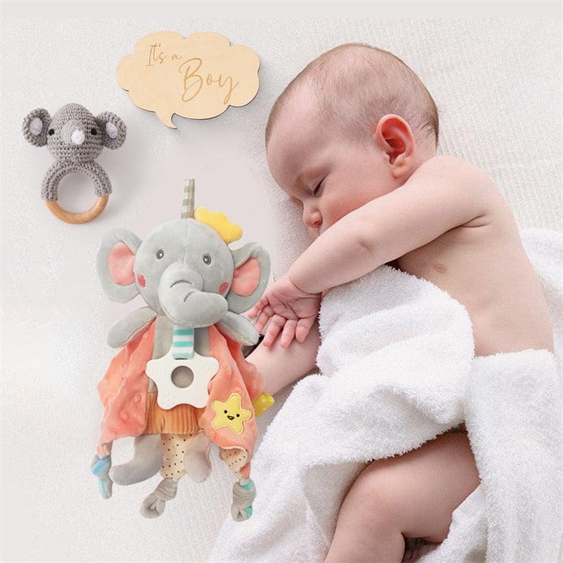 Ein in ein weißes Handtuch gewickeltes Baby schläft neben dem Ringo Baby Geschenk zur Geburt von Hello World - einem Jungen-Geschenkset mit Plüschelefant, gestrickter Koala-Rassel und hölzernem "It's a Boy"-Schild - perfekt für die Begrüßung von Neugeborenen.