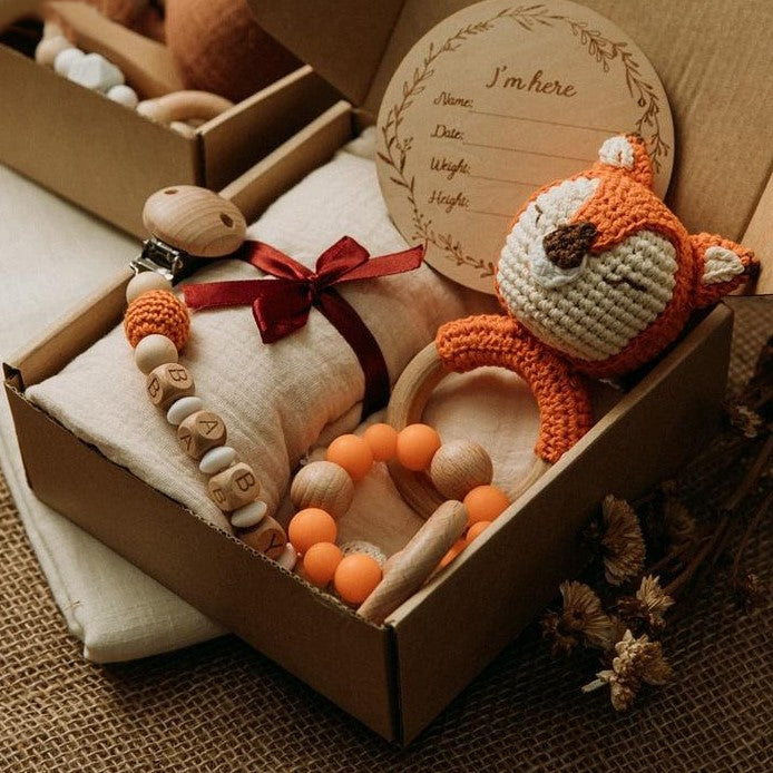 Das "Rita Baby Geschenk zur Geburt" von Hello World besteht aus einem gehäkelten orangefarbenen Fuchs, einem Holz-Silikon-Beißring, einer BABY Schnullerklemme, einem orangefarbenen Beißarmband, einer roten Schleife und einer hölzernen Geburtsanzeige auf einem weichen Kissen.