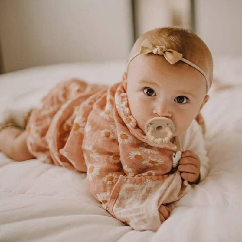 Ein Baby in einem pfirsichfarbenen Blumenkleid und einem beigen Stirnband liegt auf einem weißen Bett und blickt in die Kamera. Es hat einen Hello World Schnuller Beige [4,5 x 6 cm] Schnuller im Mund, um sich zu beruhigen.