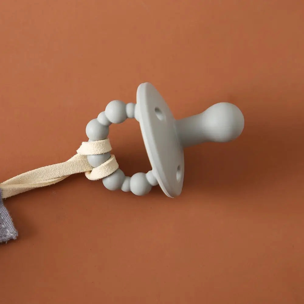 Der Hello World Schnuller Blau [4,5x6cm] hat ein weiches blaues Silikondesign mit einem Perlengriff und einem cremefarbenen Riemen, der auf einer braunen Oberfläche ruht, um Entspannung in den Tagesablauf Ihres Babys zu bringen.