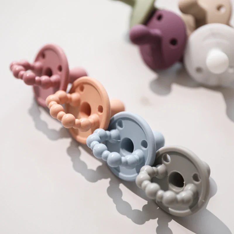 Eine diagonale Reihe von Hello World-Silikon-Babyschnullern, darunter der Schnuller Blau [4,5x6cm], erscheint in Pastellrosa, Orange, Blau und Grau auf einem hellen Hintergrund und sorgt für einen sanften Hauch von Entspannung.