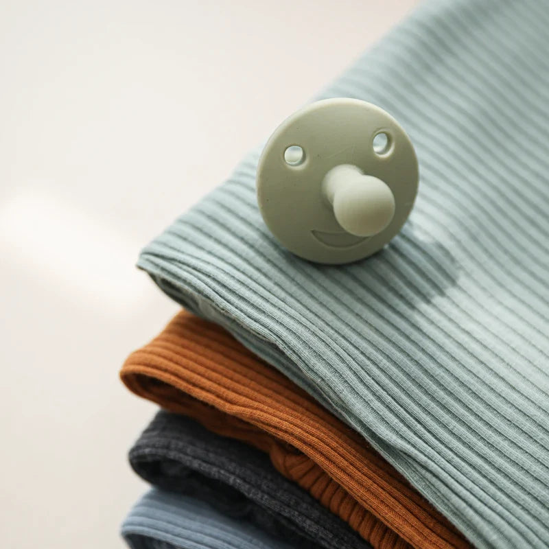 Der Schnuller Grün [4.5x6cm] von Hello World mit lächelndem Gesicht und natürlichem Design liegt auf gefalteten, gerippten Babydecken in Hellblau, Braun, Dunkelgrau und Blau.