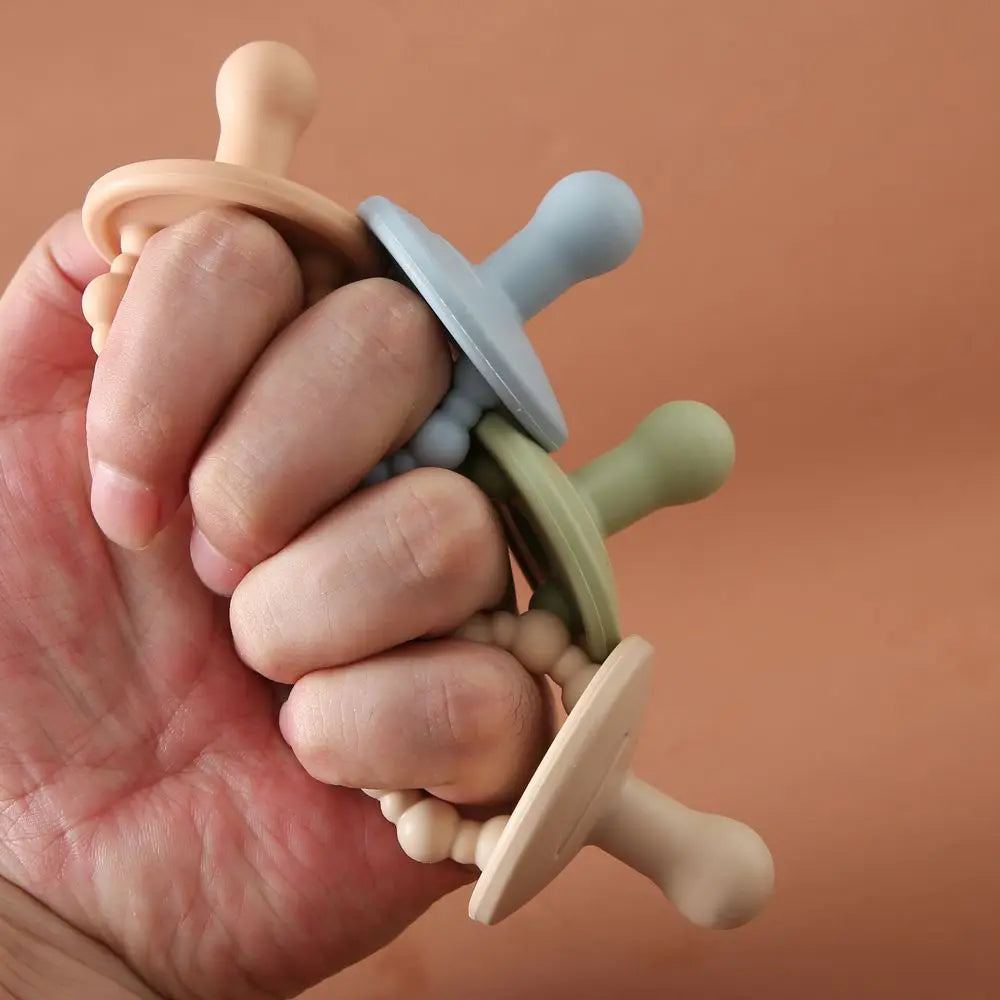 Eine Hand hält den Hello World Schnuller Grün [4,5x6cm] mit drei weiteren Mini-Schnuller in Pfirsich, Blau und Beige vor einem schlichten hellbraunen Hintergrund.