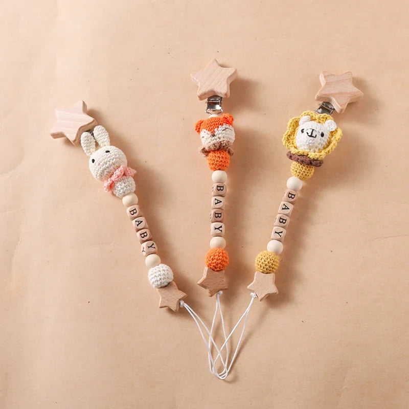 Drei Hello World Schnullerketten mit Aufschrift "Baby" Löwe [30x4,5cm] Schnullerclips mit Holzperlen und Sternclips liegen auf einer beigen Oberfläche und haben jeweils einen Holzstern an beiden Enden.