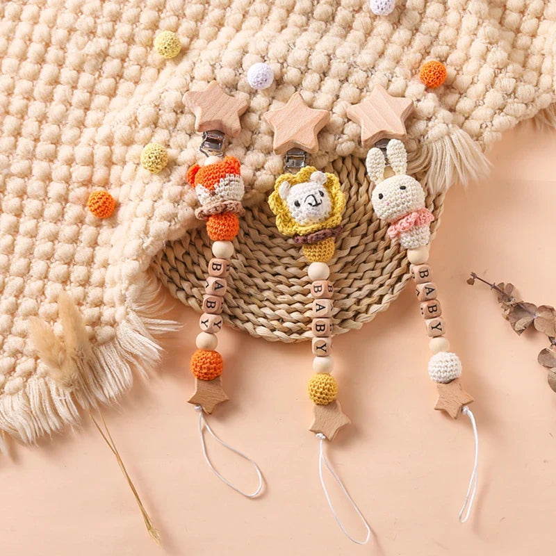 Die Hello World Schnullerkette mit Aufschrift "Baby" Löwe [30x4,5cm] besteht aus einem gehäkelten Löwen, Holzperlen und "BABY"-Buchstabenperlen, die wunderschön auf einer beigen Decke und einer gewebten Matte präsentiert werden.