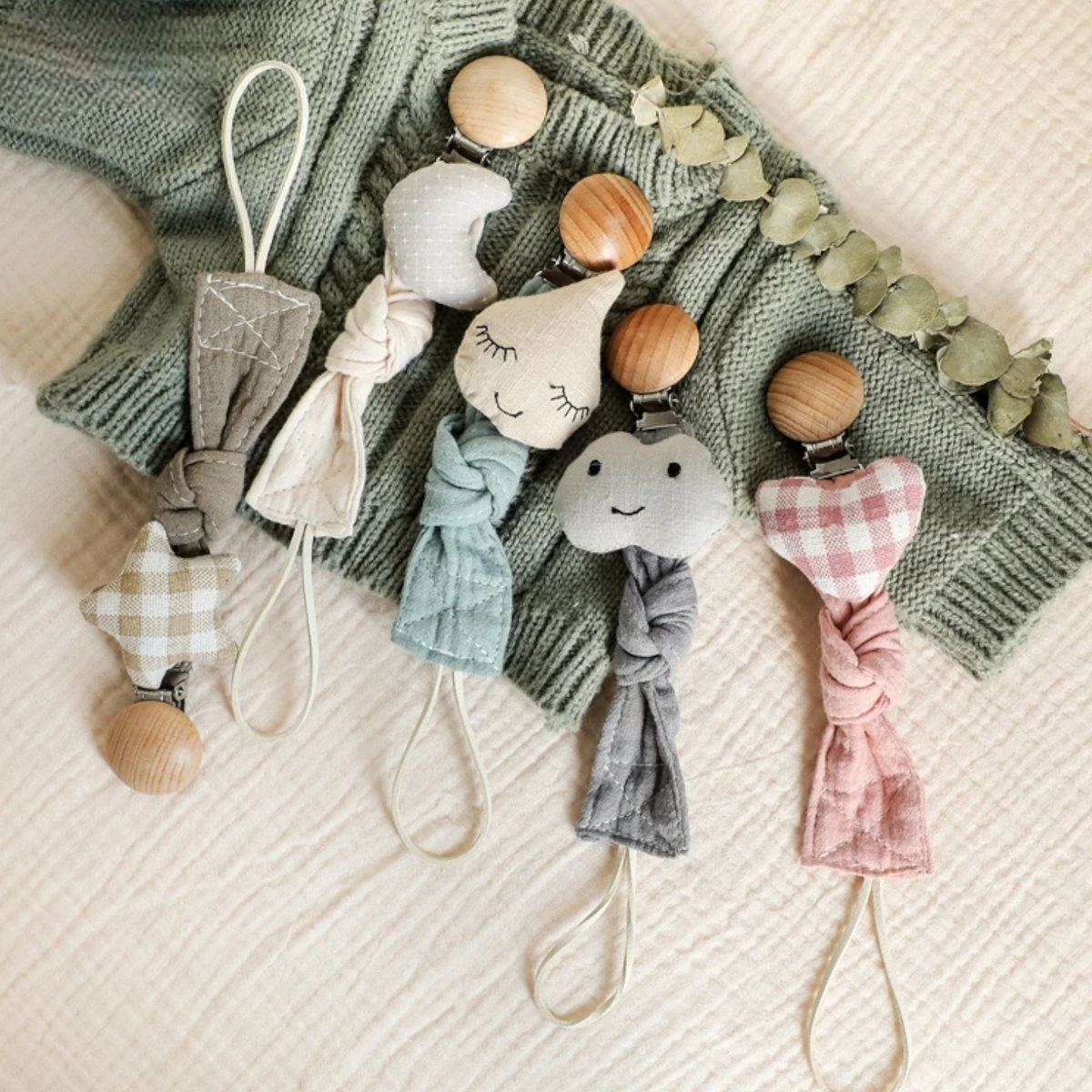 Fünf Hello World Schnullerketten mit Motiv Herz [22x3cm] mit Holzverschlüssen ruhen auf einem grünen Strickpulli und einer cremefarbenen Decke, jede mit einzigartigen Farben, Mustern und Herzmotiven für charmante Abwechslung.