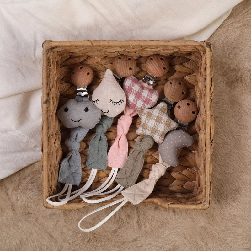 In einem geflochtenen Korb befinden sich fünf weiche Hello World Schnullerketten mit Motiv Mond [22x3cm] in verschiedenen Farben und Mustern, die auf einer plüschigen beigen Oberfläche für zusätzliche Sicherheit sorgen.