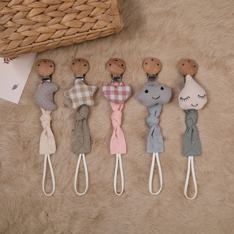Fünf "Hello World" Schnullerketten mit Regentropfen-Motiv (22x3cm) und Holzclips in Mond-, Stern-, Herz-, Wolken- und Schlafgesicht-Form liegen auf weichem Beige - für Sicherheit und Stil im Alltag.