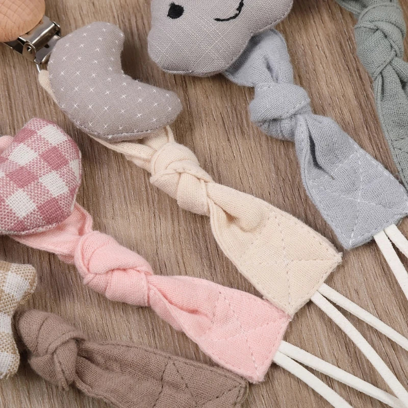 Die Schnullerkette mit Motiv Wolke [22x3cm] von Hello World ist mit einem bezaubernden Wolkendesign und einem weichen, pastellfarbenen Stoff mit Steppmuster versehen und ordentlich auf einer Holzfläche angeordnet.