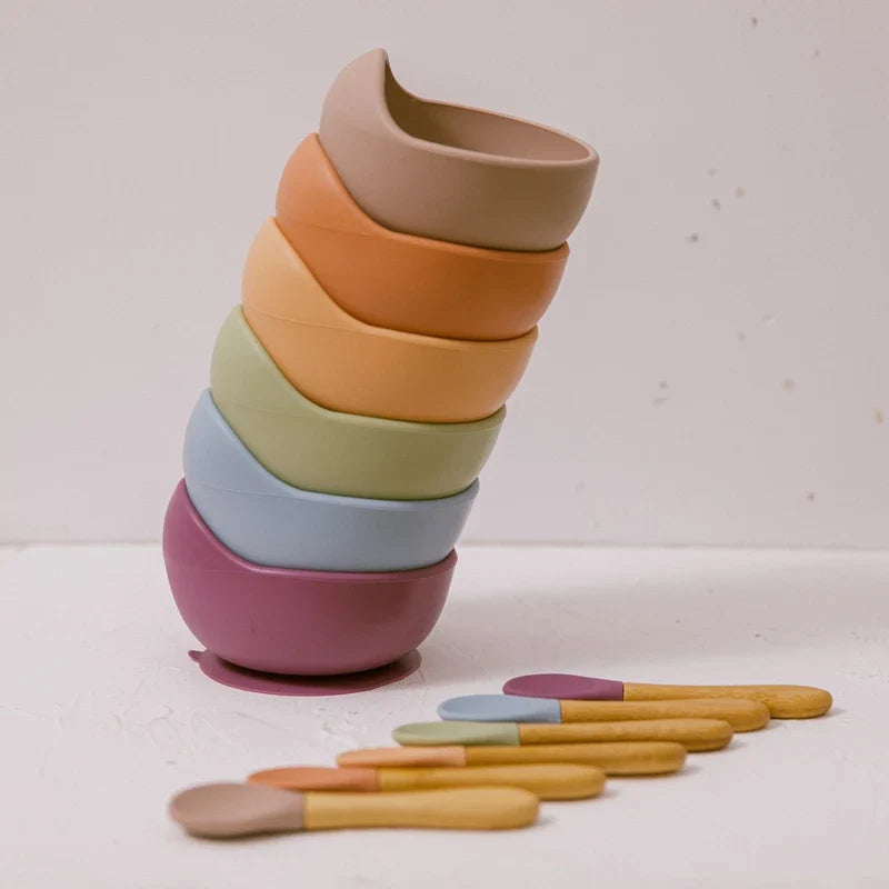 Ein Stapel Hello World Schüssel rund Beige [12x8cm] in gedeckten Tönen steht auf Weiß, mit sechs passenden Löffeln aus Silikon und Holz in einer Reihe - ideal als Kinderschüssel.