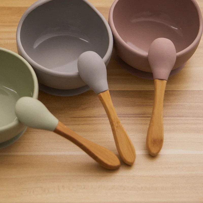 Drei BPA-freie Babylöffel mit Silikonspitzen und Bambusgriffen werden mit einer Hello World Schüssel rund Grau [12x8cm] und Schalen in Grün und Lila auf einer Holzfläche arrangiert.