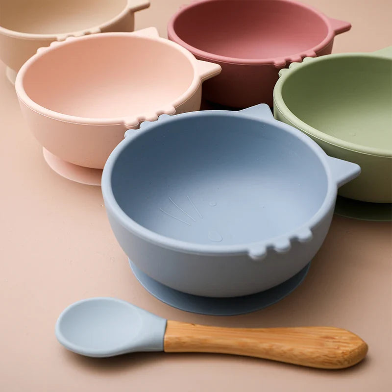 Fünf Hello World Schüssel rund Katze Beige [12x5cm] mit Katzenohren-Motiven in Pastellblau, Grün, Rosa, Pfirsich und Rose sind auf einer beigen Fläche angeordnet. Ein passender blauer Löffel mit Holzgriff vervollständigt das niedliche Set.