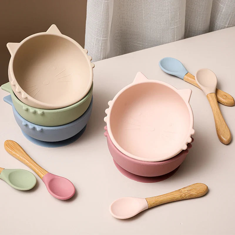 Drei Stapel von Hello Worlds Schüssel rund Katze Pink [12x5cm] mit Löffeln mit Holzgriff sind auf einer hellen Fläche angeordnet. Diese pastellrosa, grünen und blauen Schüsseln in Katzenform stehen vor einem weißen Vorhang.