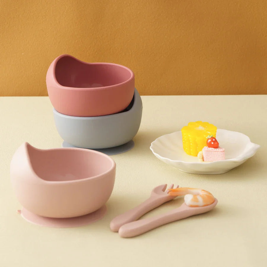 Drei Hello World Schüsseln rund Pink [12x8cm] stehen gestapelt auf einem Tisch. Davor steht ein rosafarbenes Kindergeschirr mit Schüssel, passender Gabel und Löffel sowie ein weißer Teller mit Spielzeugfutter. Der Hintergrund ist hellbraun.