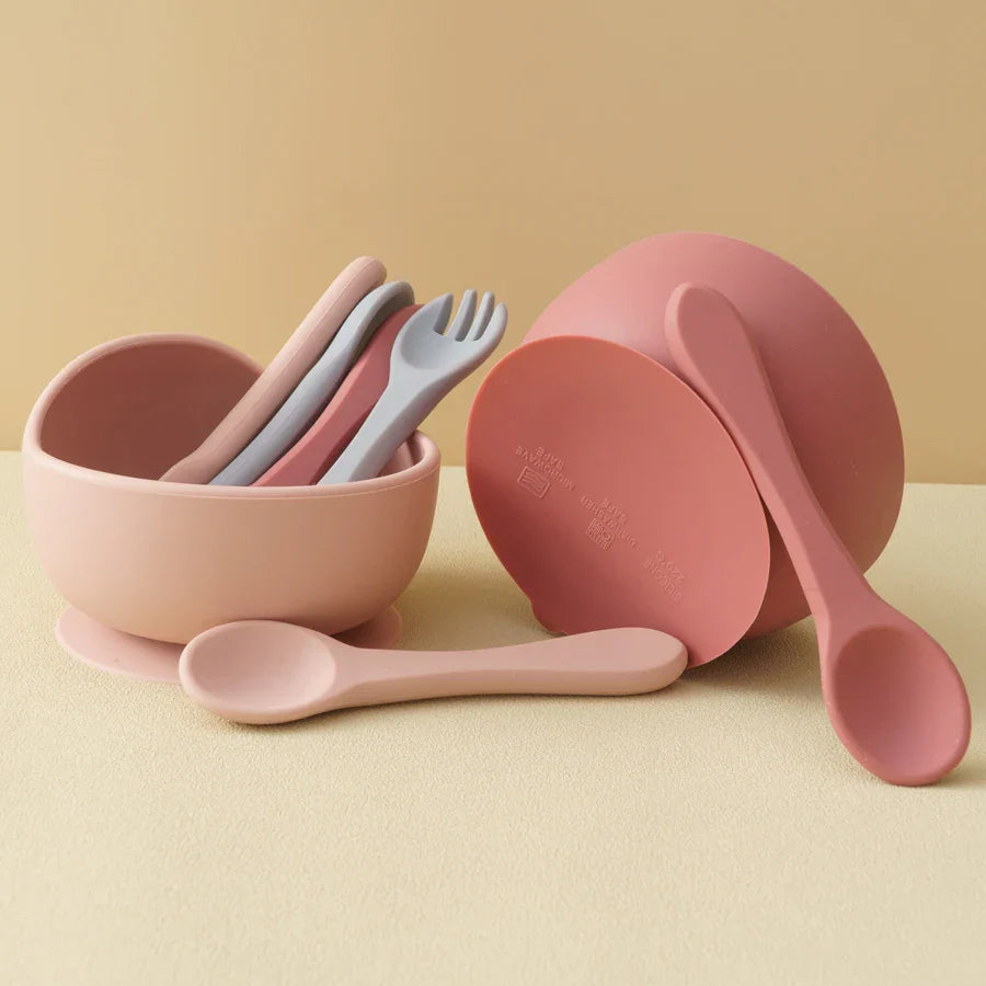 Zwei Hello World Schüsseln rund Pink [12x8cm] - eine aufrecht mit pastellfarbenen Löffeln und Gabeln, die andere umgekippt. Vorne zwei Silikonlöffel, alles auf beigem Hintergrund.