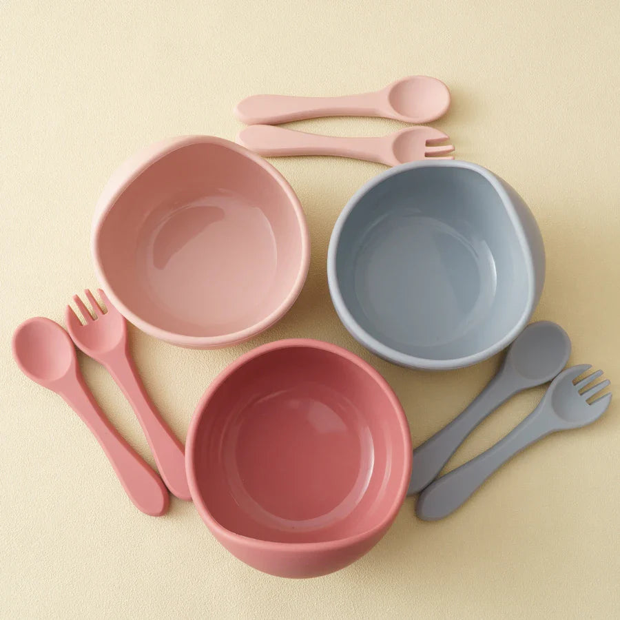 Das Hello World Schüssel rund Pink [12x8cm] Set besteht aus einer pinkfarbenen Schüssel mit passendem Löffel und Gabel, die auf einer hellbeigen Oberfläche angeordnet sind. Das minimalistische Design sorgt für glatte Utensilien für eine einfache Verwendung.