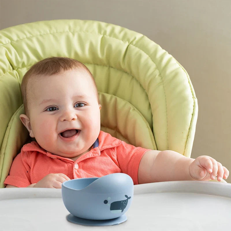 Ein lächelndes Baby in einem korallenroten Hemd sitzt in einem hellgrünen Hochstuhl und greift nach einer blauen Hello World Schüssel rund mit Motiv Blau [12x8cm] auf dem weißen Tablett.