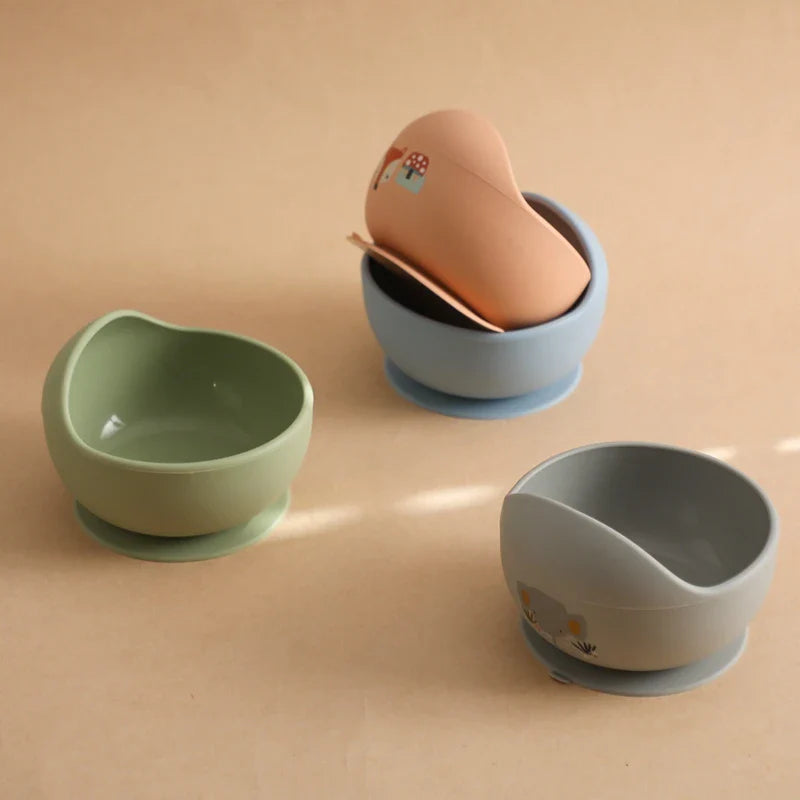 Die "Schüssel rund mit Motiv Blau [12x8cm]" von Hello World sitzt mit pastellgrünen und grauen Silikonschalen auf einer beigen Oberfläche. Die blaue Schüssel verfügt über einen pfirsichfarbenen Löffel mit Pilzmotiv, eine passende Tasse und hat einen Saugfuß für Stabilität.