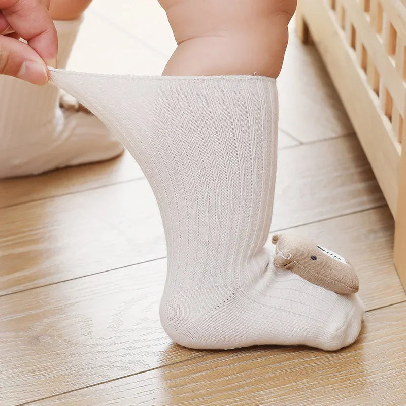 Ein Baby, das die Socken Bär (Sohle: 6x11cm, 6-12 Monate) von Hello World trägt, steht auf einem Holzboden, während eine Hand die großen, cremefarbenen Socken mit einem kleinen Bärenmuster in der Nähe der Zehen ausstreckt.