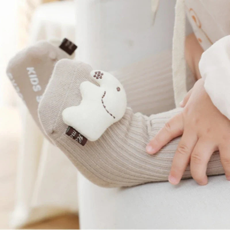 Ein Kind, das beigefarbene Hello World Socken Elefant (Sohle: 6x11cm, 6-12 Monate) mit einem lächelnden weißen Elefantenaufnäher am Knöchel trägt, sitzt auf einer hellen Fläche und berührt sanft sein Bein.