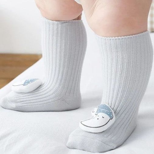 Ein Baby steht auf einer weißen Fläche und trägt Hello World Socken Mond (Sohle: 6x11cm) für 6-12 Monate - hellgraue gerippte Kniestrümpfe mit weißen und blauen lächelnden Mondmotiven an den Zehen.