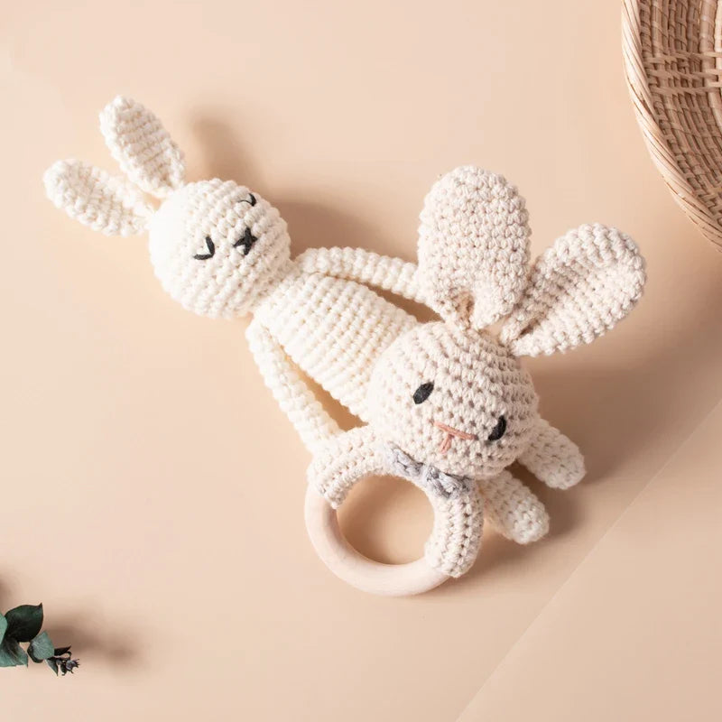 Zwei cremefarbene Hello World Stricktier Hase-Spielzeuge (18,5 x 6 cm) liegen auf einer beigen Fläche: ein Plüschhase mit geschlossenen Augen und eine lächelnde Rassel mit Holzring. In der Ecke steht ein Weidenkorb.