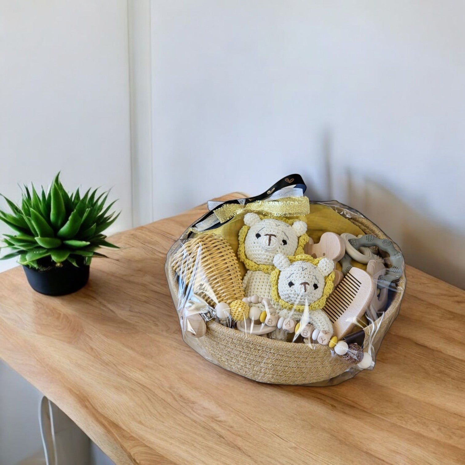 Ein mit Hello World Transparente Folie umwickelter Weidenkorb steht auf einem Holztisch, gefüllt mit Babysachen wie gehäkelten Bären, Holzbürsten, einem gelben Handtuch und Beißringen. Eine grüne Topfpflanze steht daneben.