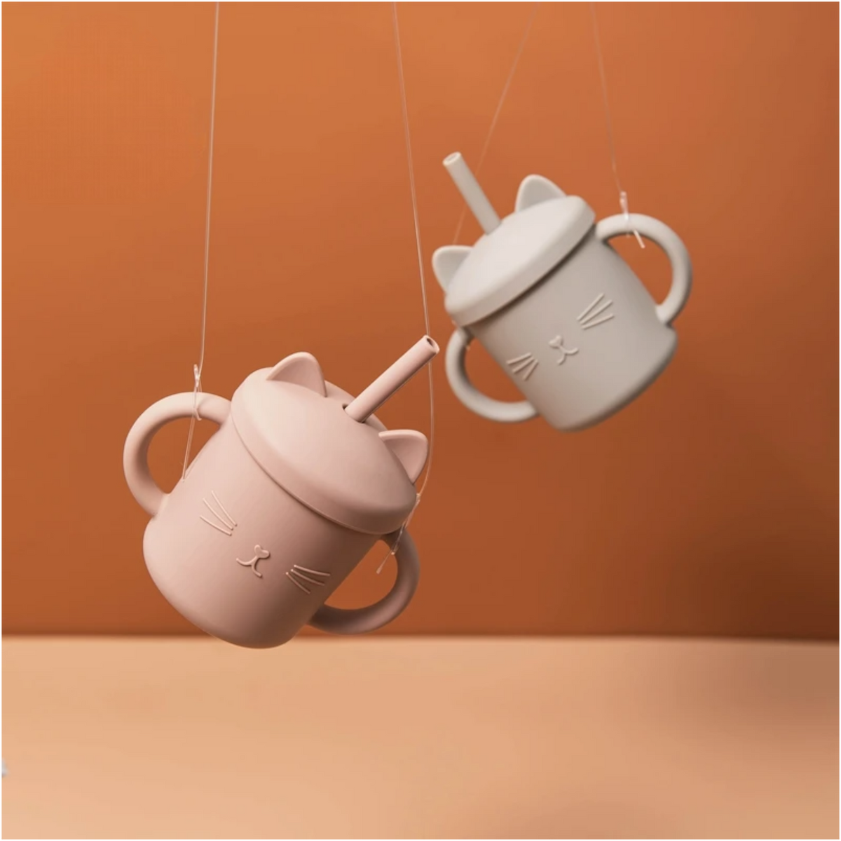 Zwei beigefarbene Hello World Trinkbecher in Katzenform (12x11cm) hängen an Schnüren vor einem unifarbenen Hintergrund. Jeder Trinkbecher hat Henkel, Strohhalme und Deckel mit Katzenohren in einem charmanten Katzen-Design.