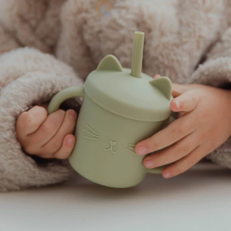 Ein Kind in einem flauschigen beigen Pullover hält den Hello World Trinkbecher Katze Grün [12x11cm] mit Katzenohren, Henkel, Strohhalm und einem niedlichen Katzengesicht auf der Vorderseite.