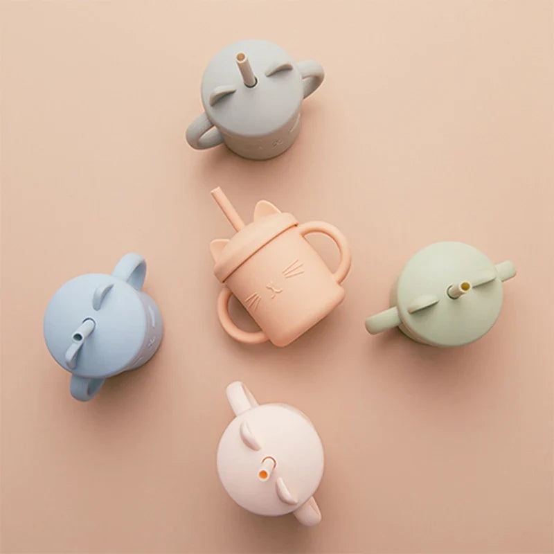 Fünf pastellfarbene Hello World Trinkbecher Katze Grün [12x11cm] mit Griffen und Deckeln sind auf einer beigen Fläche angeordnet. Eine zeigt ein Katzen-Design mit Schnurrbart, die anderen sind in den Farben Blau, Grün, Pfirsich, Rosa und Creme gehalten.