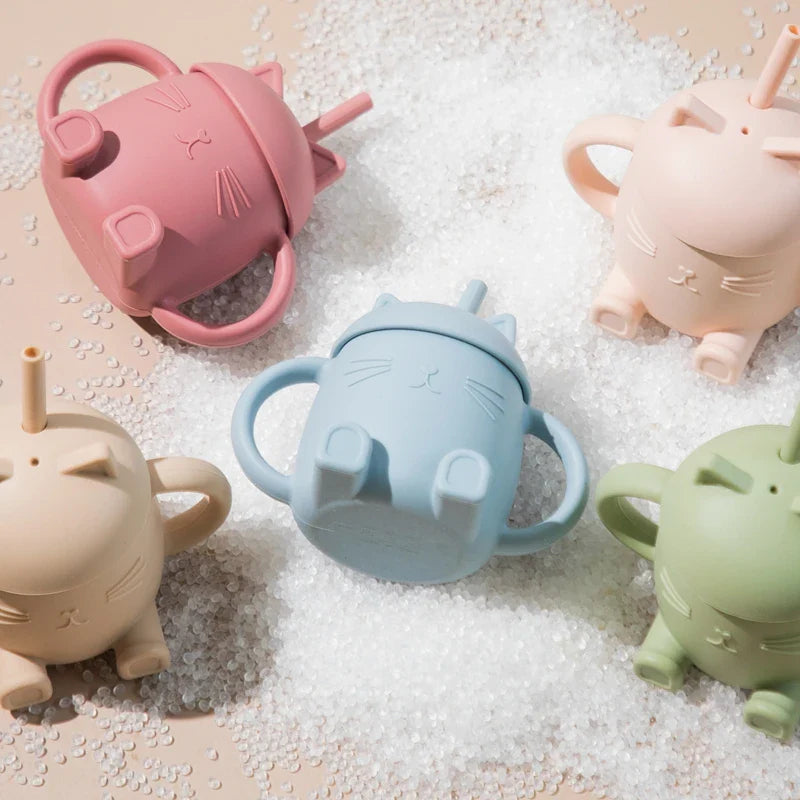 Fünf Hello World Trinkbecher Katze sitzend (13x11cm) in Pastellrosa, Blau, Beige, Grün und Blush - mit Henkel und Strohhalm - sind auf einer mit kleinen weißen Perlen bestreuten Fläche angeordnet.