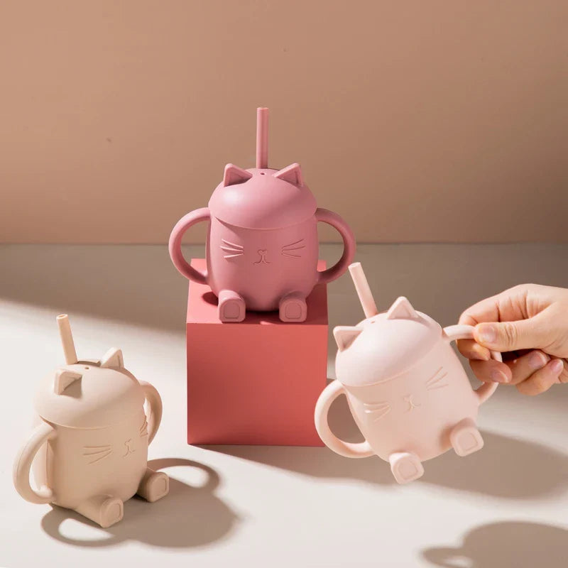 Drei Hello World Trinkbecher Katze sitzend Beige [13x11cm] mit Strohhalm und Henkel - rosa, creme und beige - sind auf einer beigen Fläche abgebildet, während einer in der Hand gehalten wird. Auf den Bechern sind süße sitzende Katzen abgebildet.