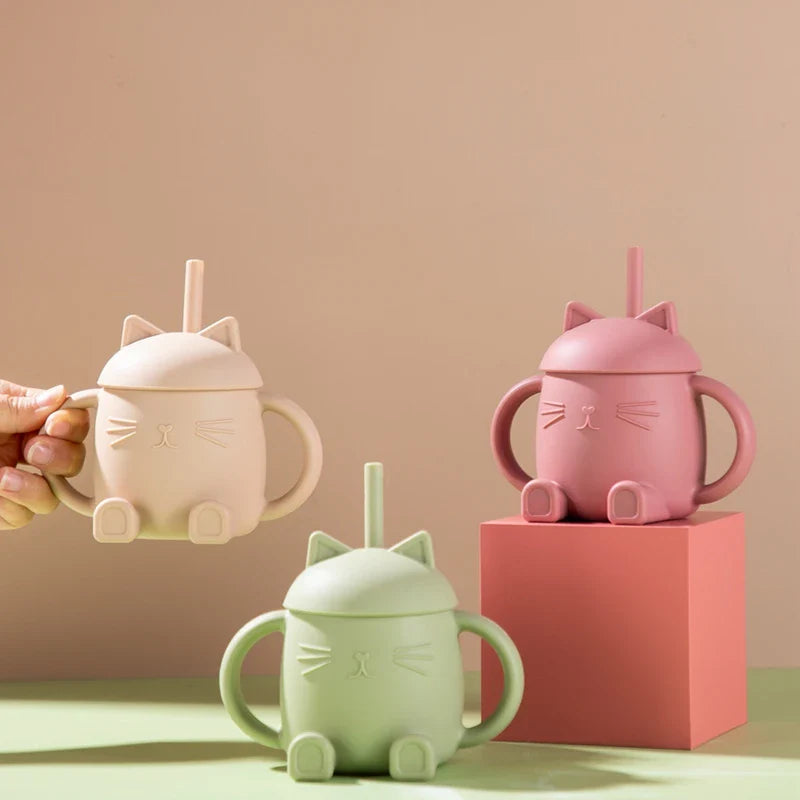 Drei Hello World Trinkbecher Katze sitzend Beige [13x11cm] mit Strohhalmen und Griffen sind abgebildet - einer gehalten, einer auf einem rosa Block und einer auf einer flachen Oberfläche. Jeder zeigt eine niedliche sitzende Katzenfigur in Beige, Rosa und Mintgrün.